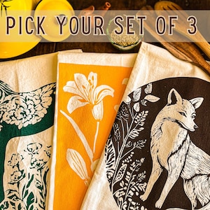 Puede incluir: Conjunto de tres toallas de cocina con diferentes diseños. Una presenta un caballo verde con flores, otra tiene un fondo amarillo con un lirio blanco, y la última tiene un fondo marrón con un zorro blanco y flores.