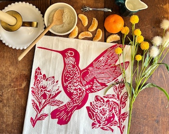 May include: A white tea towel with a pink hummingbird and floral designs printed on it. The hummingbird is flying with its wings spread out. The flowers are stylized and have a pink outline.