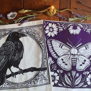 May include: Two decorative square cloths with black and white illustrations. One features a raven perched on a branch within a circle, the other a moth surrounded by flowers on a purple background. A candle holder and other decorative items are in the background.