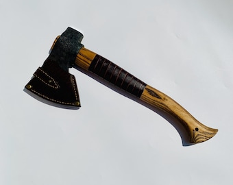 May include: A small axe with a brown leather sheath and a wooden handle with a brown leather wrap. The handle has a unique shape with a small hole at the end.