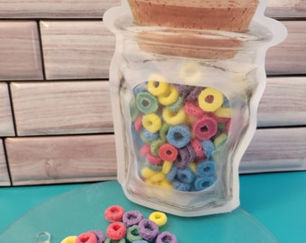 May include: A clear, jar-shaped bag filled with colorful, ring-shaped cereal. The bag has a cork-like top. Some cereal pieces are scattered on a teal-colored, round surface. The cereal pieces are red, yellow, green, blue, and purple.