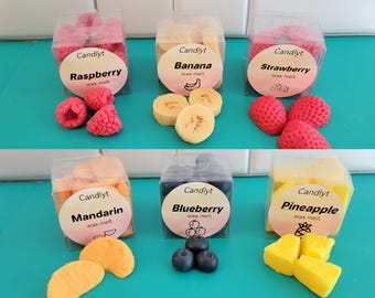 May include: Six clear plastic boxes of Candlyt wax melts, each labeled with a different fruit: raspberry, banana, strawberry, mandarin, blueberry, and pineapple. The wax melts are shaped like the fruits and are in various colors, including red, yellow, and orange.