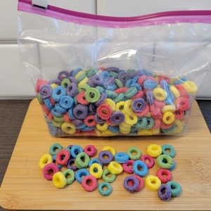 May include: A clear plastic bag filled with colorful, ring-shaped cereal. The cereal pieces are various colors, including red, yellow, blue, green, and purple. Some cereal pieces are scattered on a wooden cutting board in front of the bag.