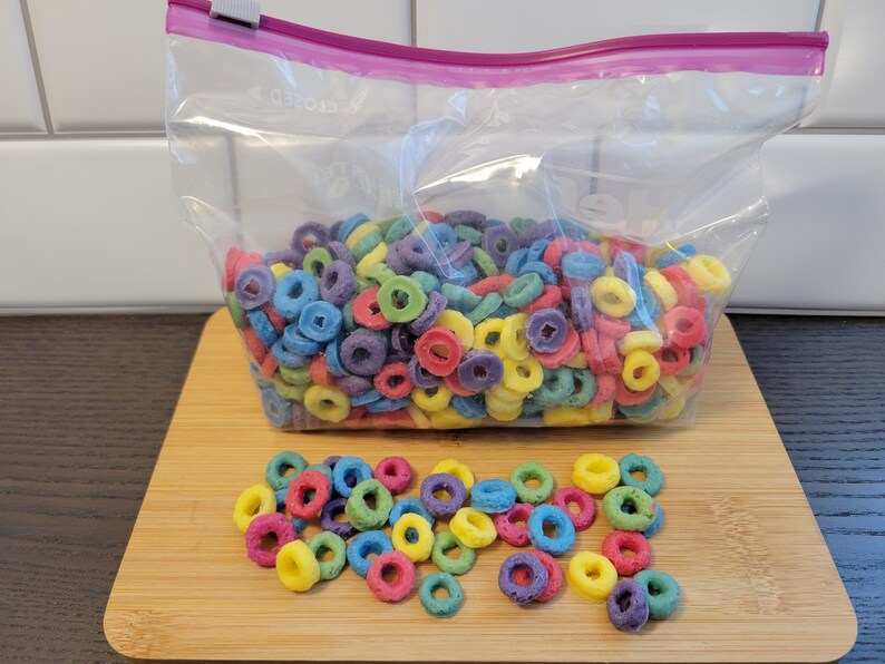 May include: A clear plastic bag filled with colorful, ring-shaped cereal. The cereal pieces are various colors, including red, yellow, blue, green, and purple. Some cereal pieces are scattered on a wooden cutting board in front of the bag.