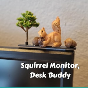 May include: A brown squirrel figurine holding a dark nut, positioned on a black rectangular base with a miniature green tree and two brown acorns. The text "Squirrel Monitor, Desk Buddy" is printed on the front of the black monitor frame.