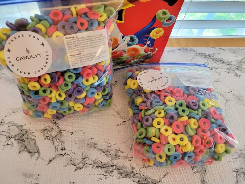 May include: Two clear plastic bags filled with colorful, ring-shaped wax melts. Each bag has a white sticker with the text "THANK YOU FOR YOUR PURCHASE" and the brand name "CANDLYT". A cereal box is visible in the background.