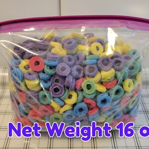 May include: A clear plastic bag filled with colorful, ring-shaped cereal. The cereal pieces are in various colors, including red, purple, yellow, and blue. The bag is sealed with a purple closure and has the text "Net Weight 16 oz".