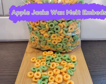 May include: A bag of green and orange cereal rings labeled "Apple Jacks Wax Melt Embeds". The cereal is on a wooden cutting board.