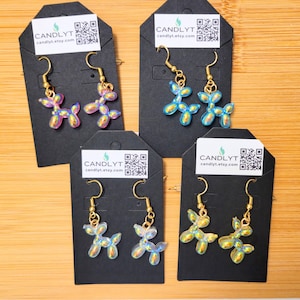 May include: Four pairs of dog-shaped earrings with gold-colored hooks. The earrings are iridescent, with colors shifting from pink to blue and yellow. Each pair is displayed on a black card with the text "CANDLYT" and a QR code.