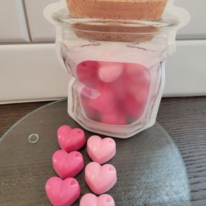 May include: Six pink heart-shaped wax melts in various shades of pink, arranged in a row on a glass surface. A clear plastic bag with a cork lid and pink wax melts inside is in the background.