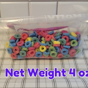 May include: A clear plastic bag filled with colorful, ring-shaped cereal. The cereal pieces are in various colors, including red, blue, yellow, and green. The text "Net Weight 4 oz" is displayed in a purple font.