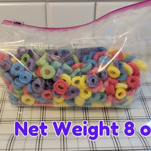 May include: A clear plastic bag filled with colorful, ring-shaped cereal. The cereal pieces are in various colors, including red, yellow, green, blue, and purple. The bag is sealed with a pink zipper and the text "Net Weight 8 oz" is visible.