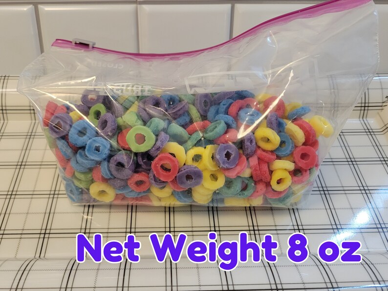 May include: A clear plastic bag filled with colorful, ring-shaped cereal. The cereal pieces are in various colors, including red, yellow, green, blue, and purple. The bag is sealed with a pink zipper and the text "Net Weight 8 oz" is visible.