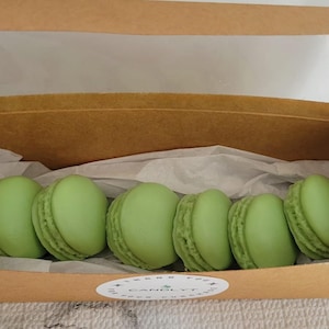 May include: A box of six green macarons in a clear plastic window box. The box is brown and has a white label with the text "CANDLY T" on it.