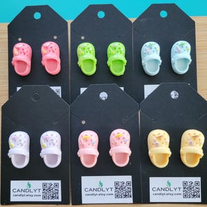 May include: Six pairs of miniature clog earrings in various colors, including pink, green, blue, white, and yellow. Each pair is displayed on a black card with the shop's name and a QR code. The earrings are a playful accessory.