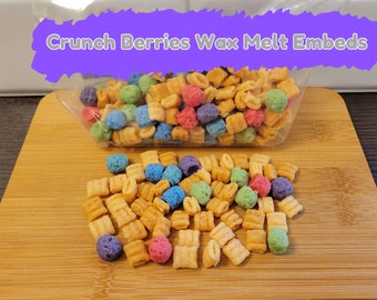 May include: A plastic bag filled with colorful cereal squares and round cereal pieces. The bag is labeled "Crunch Berries Wax Melt Embeds". The cereal is scattered on a wooden surface.