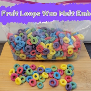 May include: A clear plastic bag filled with colorful, ring-shaped wax melt embeds, resembling Fruit Loops cereal. The bag sits on a wooden cutting board, with a small pile of the embeds scattered in front. Text on the bag reads "Fruit Loops Wax Melt Embeds."