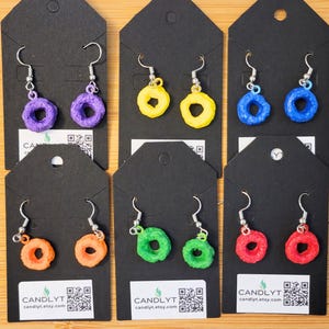 May include: A collection of colorful, handmade earrings shaped like cereal rings. The earrings are displayed on black cards with silver hooks. Colors include purple, yellow, blue, orange, green, and red. The shop name and website are printed on the cards.