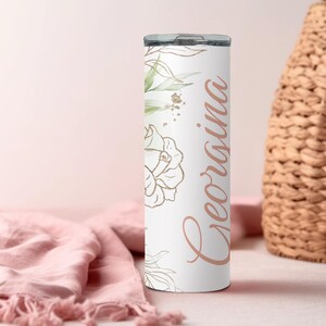 May include: A white tumbler with a floral design and the name "Georgina" in a cursive font. The tumbler features gold and green botanical accents, including leaves and flowers. The design is printed on a stainless steel insulated tumbler.