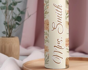 May include: A stainless steel tumbler with a floral design and the name "Mrs. Smith" in a brown script. The tumbler has a cream-colored background with peach-colored flowers and green leaves.