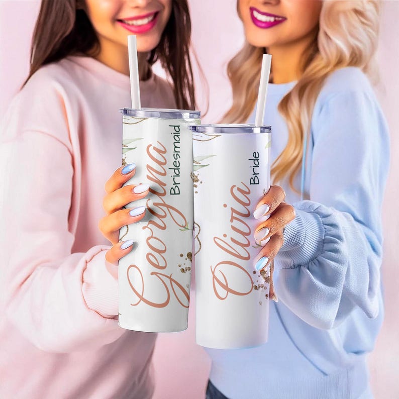 May include: Two white stainless steel tumblers with straws are held by two people. One tumbler says "Georgina Bridesmaid" in a copper script font, and the other says "Olivia Bride". Both tumblers have a floral design.