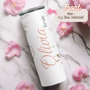 May include: White tumbler with a floral design and the name "Olivia Bride" in a soft pink script. The tumbler has a silver lid and is decorated with gold glitter accents and green leaves. The background features pink flower petals.