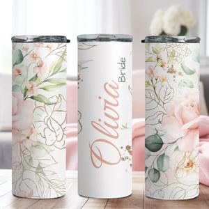 May include: White insulated tumbler with a floral design featuring pink roses, green leaves, and gold accents. The tumbler has a black lid and the name "Olivia Bride" in a rose gold script. A decorative item for a wedding.