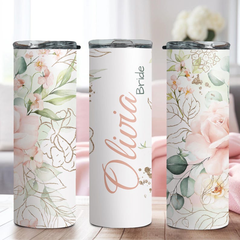 May include: White insulated tumbler with a floral design featuring pink roses, green leaves, and gold accents. The tumbler has a black lid and the name "Olivia Bride" in a rose gold script. A decorative item for a wedding.