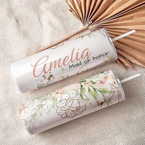 May include: Two white insulated tumblers with floral designs and straws. One tumbler has the name "Amelia" and "Maid of honor" in a script font. The other tumbler has a floral design with gold accents and green leaves.