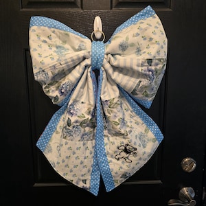 Könnte beinhalten: Eine große, dekorative Schleife aus Stoff mit einem Patchwork-Design aus Blumen- und Streifenmustern in Blau- und Weißtönen. Die Schleife hat einen blauen Polka-Dot-Rand und ein gesticktes Bärenemblem. Sie hängt an einer schwarzen Tür.