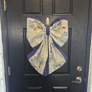May include: A fabric bow decoration, suspended on a black door. The bow has a floral design with blue hydrangeas on a pale backdrop, contrasted with navy blue polka dots and gingham patterns.