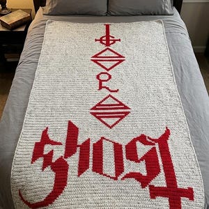 Könnte beinhalten: Eine gehäkelte Decke in Hellgrau mit roten Buchstaben und Symbolen. Das Wort "GHOST" ist in einer gotischen Schriftart dargestellt. Die rechteckige Decke liegt auf einem Bett.