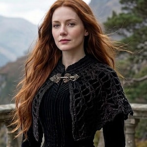 May include: A black crocheted capelet featuring an open-work lattice pattern and a decorative metal clasp at the neckline, worn over a black velvet long-sleeved garment with vertical ribbed detailing on the bodice.
