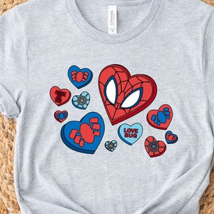May include: A gray t-shirt with a graphic of a red and blue heart with a spider-man face on it. The heart is surrounded by smaller hearts with different designs. The text "Love Bug" is on one of the smaller hearts.