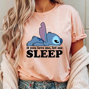 Puede incluir: Una camiseta rosa con un personaje de dibujos animados azul de Stitch de Lilo & Stitch acostado. El texto "if you love me, let me SLEEP" est&aacute; impreso en negro debajo del personaje.