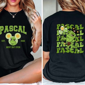 May include: Black t-shirt with a green chameleon cartoon character wearing a Mickey Mouse hat and holding balloons. The text "PASCAL" is printed on the shirt in a retro font with the words "EST. 2010" and "BEST DAY EVER" printed below the character.