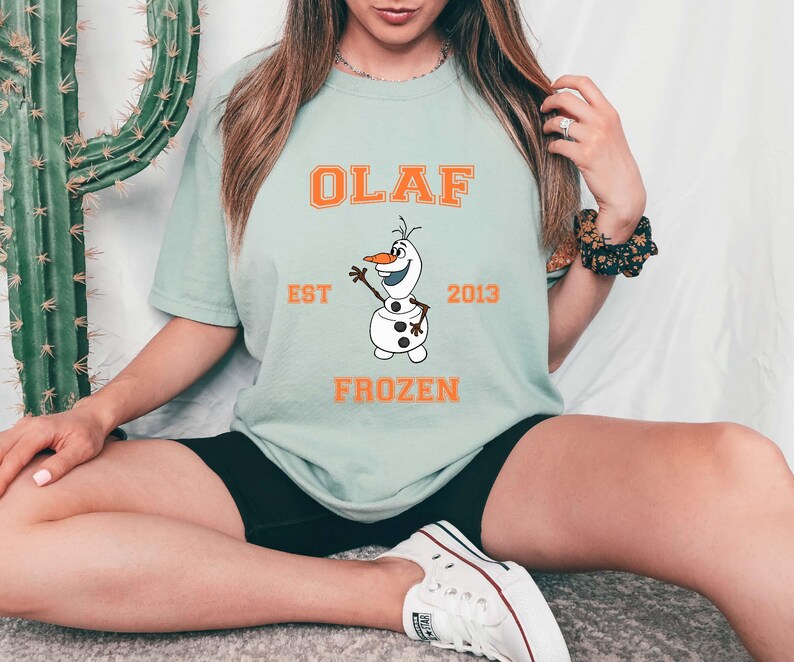 Puede incluir: Una camiseta azul claro con una imagen de dibujos animados de Olaf de Frozen. La camiseta tiene texto naranja que dice "Olaf Est 2013 Frozen".