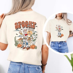Puede incluir: Una camiseta beige con un dibujo animado de una momia vestida de vaquero, sosteniendo una pistola. El texto "Spooky Vibes" est&aacute; por encima de la momia. La camiseta est&aacute; siendo modelada por una persona que lleva pantalones vaqueros azules.