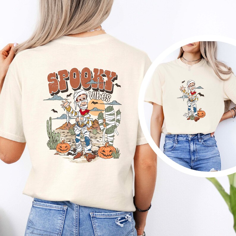 Puede incluir: Una camiseta beige con un dibujo animado de una momia vestida de vaquero, sosteniendo una pistola. El texto "Spooky Vibes" est&aacute; por encima de la momia. La camiseta est&aacute; siendo modelada por una persona que lleva pantalones vaqueros azules.