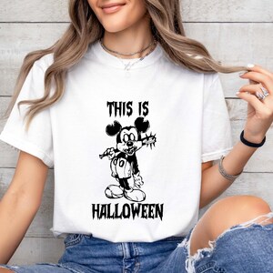May include: White t-shirt featuring a black and white graphic of a cartoon mouse holding a weapon, with the text "THIS IS HALLOWEEN" in a gothic font. The shirt is a short-sleeved crew neck.