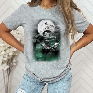 K&ouml;nnte beinhalten: Ein graues T-Shirt mit einem Grafikdruck von Figuren aus dem Film Nightmare Before Christmas. Die Grafik zeigt Jack Skellington, Sally und Oogie Boogie vor einem Vollmond.