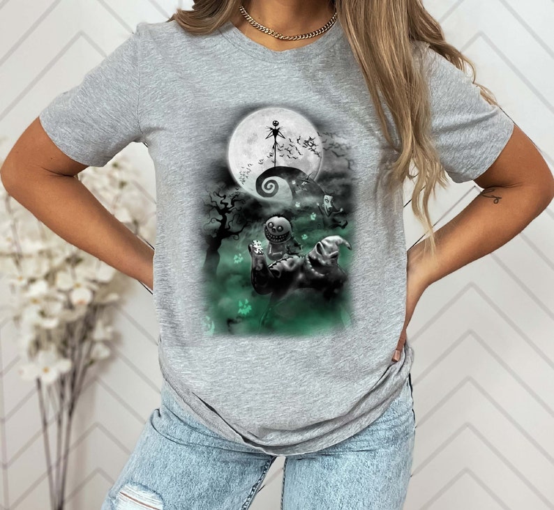 K&ouml;nnte beinhalten: Ein graues T-Shirt mit einem Grafikdruck von Figuren aus dem Film Nightmare Before Christmas. Die Grafik zeigt Jack Skellington, Sally und Oogie Boogie vor einem Vollmond.