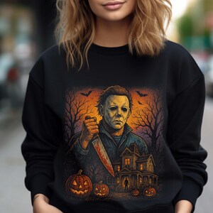 Może przedstawiać: Czarna bluza z nadrukiem o tematyce Halloween. Grafika przedstawia groźną postać trzymającą zakrwawiony n&oacute;ż, nawiedzony dom, dynie i nietoperze na upiornym pomarańczowo-fioletowym tle. Sezonowy element odzieży.