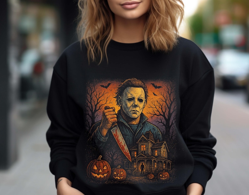 Może przedstawiać: Czarna bluza z nadrukiem o tematyce Halloween. Grafika przedstawia groźną postać trzymającą zakrwawiony n&oacute;ż, nawiedzony dom, dynie i nietoperze na upiornym pomarańczowo-fioletowym tle. Sezonowy element odzieży.