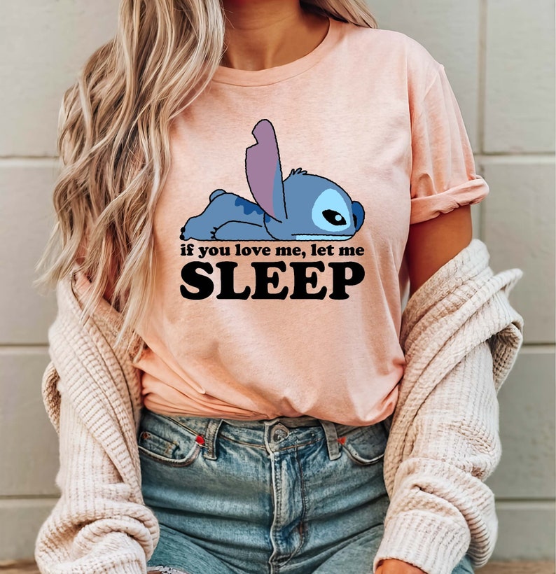 Puede incluir: Una camiseta rosa con un personaje de dibujos animados azul de Stitch de Lilo & Stitch acostado. El texto "if you love me, let me SLEEP" est&aacute; impreso en negro debajo del personaje.