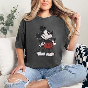 May include: Dark gray t-shirt featuring a distressed graphic of Mickey Mouse. The cartoon character is depicted in black, white, and red. The shirt has short sleeves and a relaxed fit, suitable for casual wear.