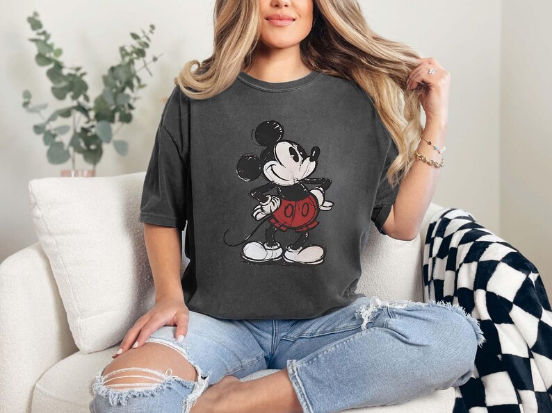 May include: Dark gray t-shirt featuring a distressed graphic of Mickey Mouse. The cartoon character is depicted in black, white, and red. The shirt has short sleeves and a relaxed fit, suitable for casual wear.
