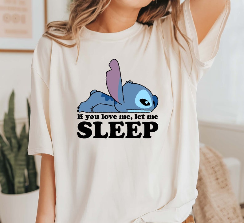 Puede incluir: Una camiseta blanca con un personaje de dibujos animados azul acostado y el texto "if you love me, let me SLEEP".