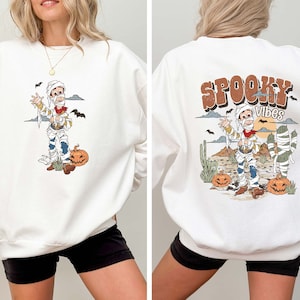 Puede incluir: Sudadera blanca con un dibujo animado de una momia con un sombrero de vaquero y sosteniendo una pistola. La parte delantera de la sudadera tiene un gr&aacute;fico de la momia en un entorno des&eacute;rtico. La parte trasera de la sudadera tiene el texto "SPOOKY VIBES" en letras marrones con un dibujo animado de una momia en un entorno des&eacute;rtico.