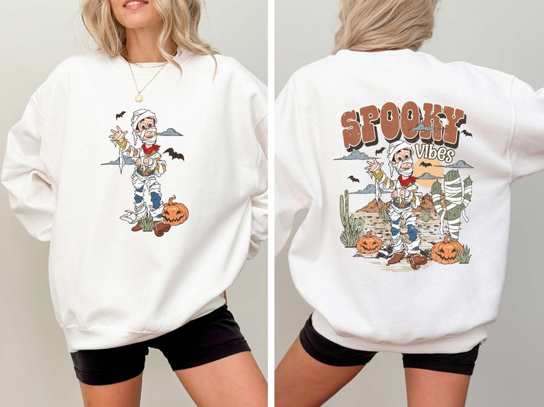 Puede incluir: Sudadera blanca con un dibujo animado de una momia con un sombrero de vaquero y sosteniendo una pistola. La parte delantera de la sudadera tiene un gr&aacute;fico de la momia en un entorno des&eacute;rtico. La parte trasera de la sudadera tiene el texto "SPOOKY VIBES" en letras marrones con un dibujo animado de una momia en un entorno des&eacute;rtico.